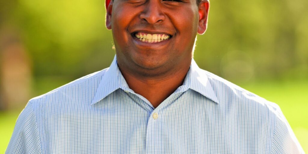 This is an image of Joe Neguse, candidate for Colorado's 2nd Congressional District. He is pictured in a blue shirt standing with a blurred background of trees.