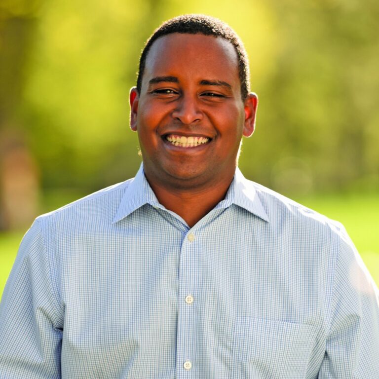 This is an image of Joe Neguse, candidate for Colorado's 2nd Congressional District. He is pictured in a blue shirt standing with a blurred background of trees.
