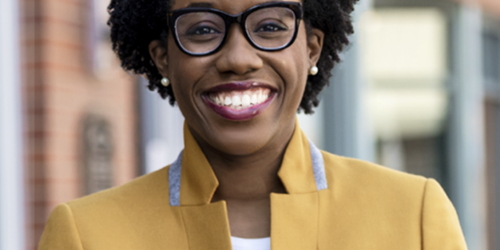 This is an image of Lauren Underwood, candidate for Illinois's 14th Congressional District. She is pictured in a dark yellow jacket in front of a buidling.
