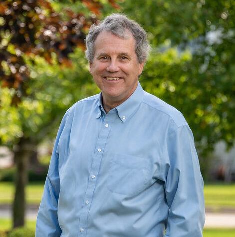 This is an image of Sherrod Brown, candidate for Ohio Senate. He is pictured in a blue shirt standing in front of trees in front of a road.