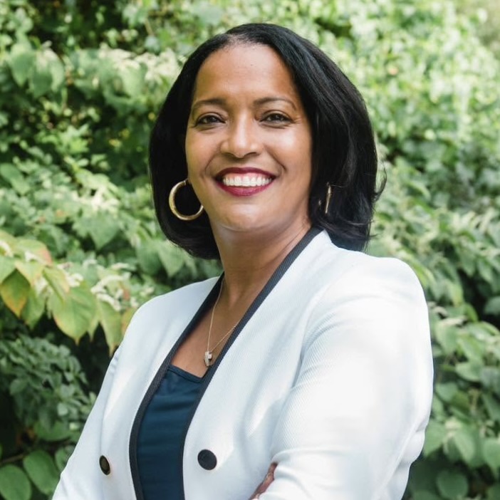 This is an image of Jahana Hayes, candidate for Connecticut's 5th Congressional District. She is pictured in a white jacket with a blue shirt standing outside in front greenery.