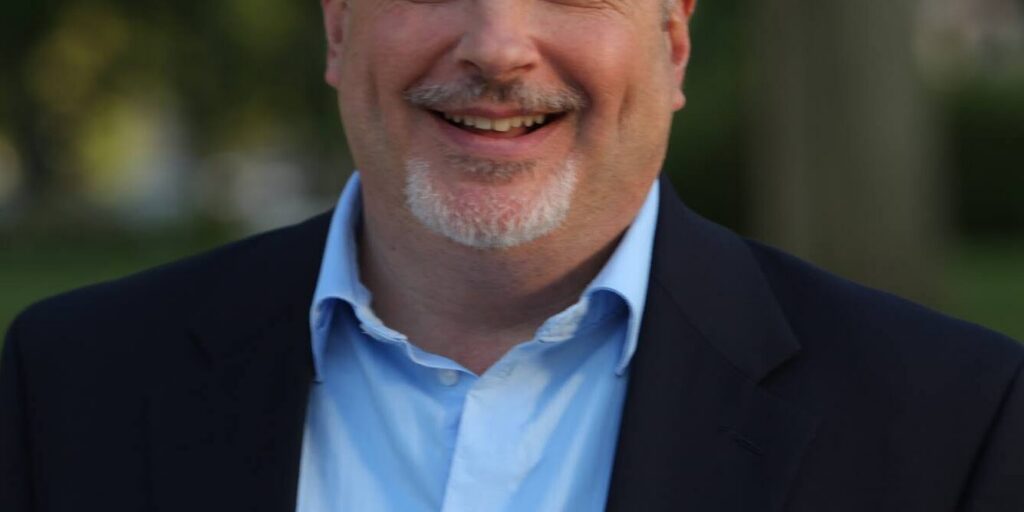 This is an image of Mark Pocan, candidate for Wisconsin's 2nd Congressional District. He is pictured in a black suit with a light blue shirt standing in front a tree.