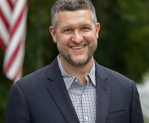 This is an image of Pat Ryan, candidate for New York's 18th Congressional District. He is pictured in a gray suit with a blue shirt standing outside in front of the American flag.