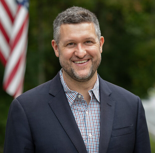 This is an image of Pat Ryan, candidate for New York's 18th Congressional District. He is pictured in a gray suit with a blue shirt standing outside in front of the American flag.