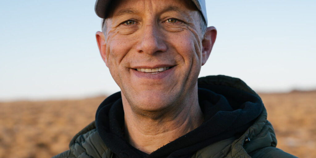 This is an image of Bill Hill, candidate for Alaska's At-Large Congressional District. He is pictured in a green jacket with a black shirt and gray hat standing on a field.