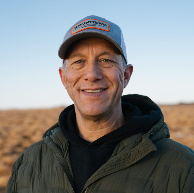 This is an image of Bill Hill, candidate for Alaska's At-Large Congressional District. He is pictured in a green jacket with a black shirt and gray hat standing on a field.