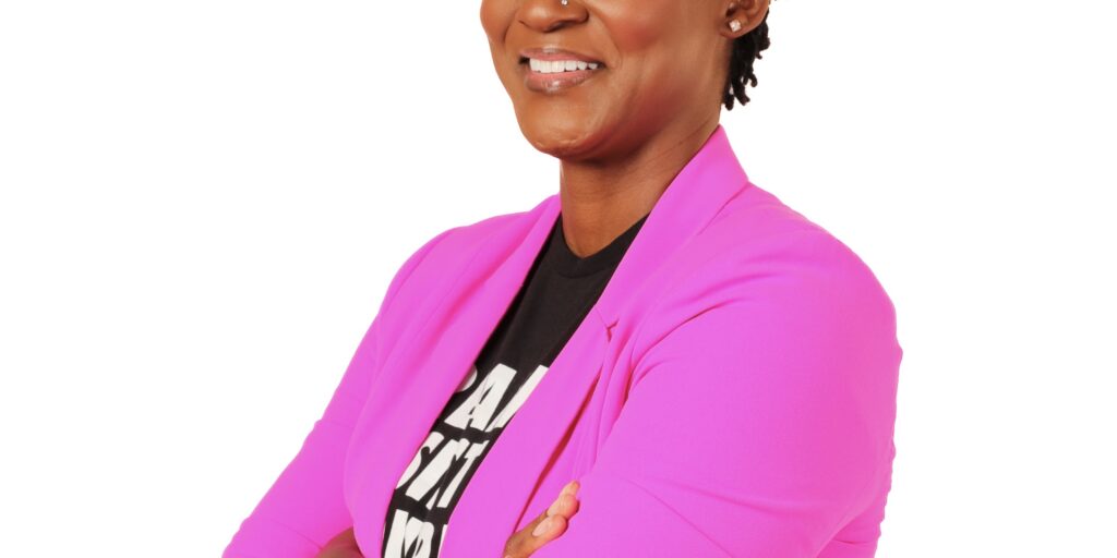 This is an image of Angie Nixon, candidate for Florida Senate. She is pictured in a pink suit with a black shirt with her arms crossed in front of a white background.